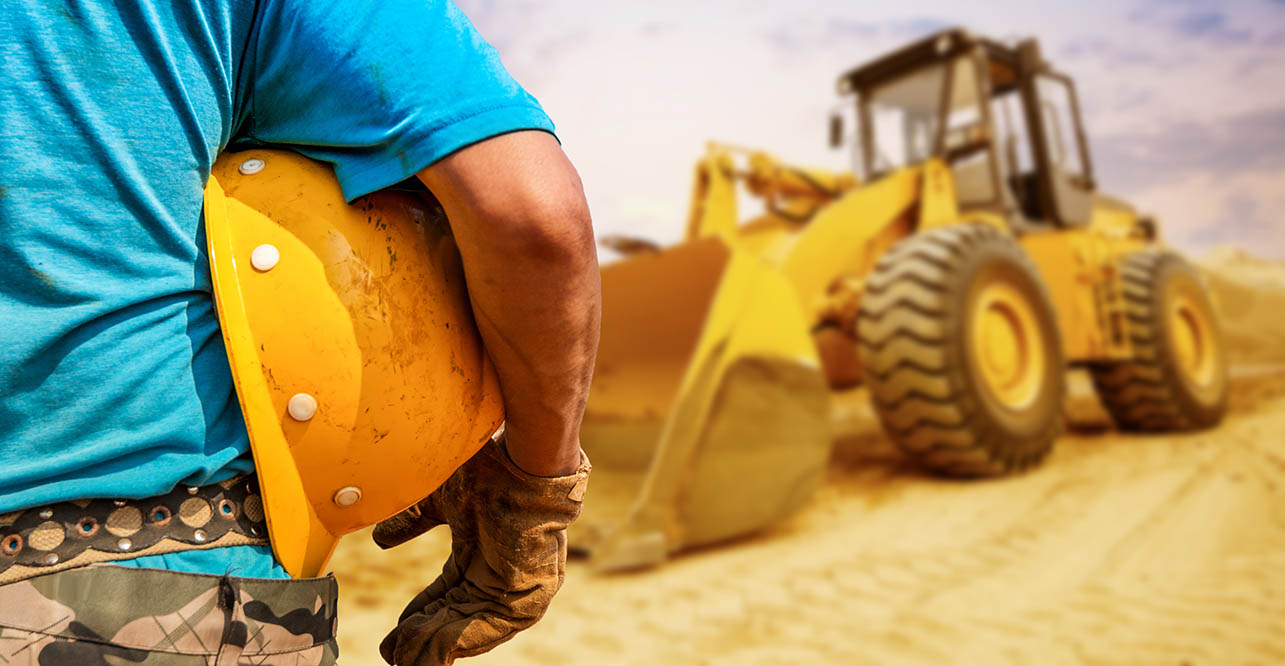 a construction worker holding a helmet, with a heavy piece of equipment in the background